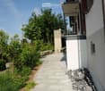 A well-maintained garden area with a paved pathway leading to a modern building. The garden features neatly trimmed bushes, small trees, and a stone wall. A balcony with a metal railing is visible on the building.