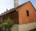 Two-story house with wooden siding and red-tiled roof, featuring multiple chimneys and windows.