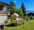 A well-maintained garden with a wooden playhouse, slide, swing, and benches, surrounded by trees and a view of mountains.