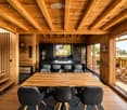 Open-concept living and dining area with exposed wooden beams, a wooden dining table with six chairs, a kitchen island with four stools, and a view to a wooden deck with outdoor seating.