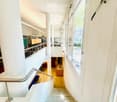 Modern interior staircase with white walls, large windows, metal handrail, and visible furniture in the background including chairs and a table.