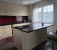 Modern kitchen with light wood cabinets, black granite countertop, sink, and a small island with a stool. Large windows with sheer curtains provide natural light.