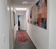 Clean, modern hallway with grey tiled floor, white walls, and a colorful abstract quilt hanging on one wall. A patterned rug lies in the center, and several doors are visible along the corridor.