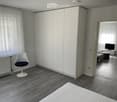 Modern furnished room with a white wardrobe, a sleek white chair, a small table, and a TV screen in the adjacent area.