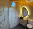 Modern bathroom with a glass-enclosed shower, a vanity with a sink and mirror, and a toilet. The room features dark tiled floors and a skylight above the shower area.