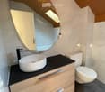 Modern bathroom with a sleek wooden vanity, black countertop, white sink, and a round mirror above it. The toilet has a decorative sticker on the lid.