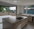 Modern kitchen with a central island featuring an induction cooktop, built-in cabinets, and a sink with a modern faucet. Large windows and a sliding glass door provide natural light and access to the outdoor area.