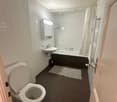 Modern bathroom with white tiles, a toilet, a sink, a bathtub, a shower, and a mirror. The floor is dark grey, and there is a small rug in front of the bathtub.