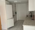 A small kitchen with white tiled flooring, white cabinetry, and a granite countertop. It includes an oven and a stove, a sink with a faucet, and a small area for a dishwasher.