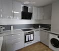 Modern kitchen with grey cabinets, white countertops, and stainless steel appliances including a microwave, oven, washing machine, coffee maker, and toaster. Wooden flooring and a sleek design.