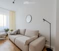 Modern living room with a light grey sectional sofa, matching armchair, floor lamp, round wall mirror, and small potted plants. The room features light wooden flooring and large windows with sheer curtains.