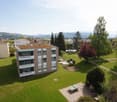 Multi-story residential building with balconies and a green lawn area in front, surrounded by trees and a scenic mountain backdrop.