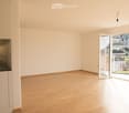 Empty room with light wooden flooring, white walls, and a small kitchen area with a sink and faucet. A sliding glass door opens to a balcony with a view of the city.
