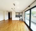 Bright, unfurnished living space with wooden flooring, large glass sliding doors leading to a balcony, and a modern kitchen area visible in the background.