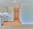 Bright, modern hallway with light wooden flooring, white walls, and recessed lighting. The hallway leads to a kitchen area with white cabinets, a countertop, and a sink.