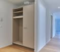 Empty hallway with light wooden flooring, white walls, built-in closet with open shelving, and recessed ceiling lighting.