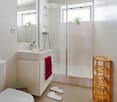 Modern bathroom with white tiles, a sink, mirror, toilet, glass-enclosed shower, a red towel, a wooden shelf, and two white bathroom slippers on the floor.