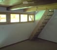 An attic space with exposed wooden beams, a wooden staircase leading up, and several windows letting in natural light. The room is empty with no furniture or furnishings.