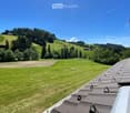 A panoramic view of a lush green valley with rolling hills, scattered trees, and a few houses in the distance under a clear blue sky.