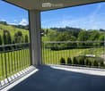 Balcony with metal railing, tiled floor, overlooking green hills and trees under a clear blue sky.