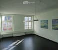 Empty room with bare walls, wooden floor, large windows, and several paintings on the walls.
