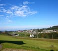 A panoramic view of a rural village nestled in a green valley, with scattered houses, trees, and open fields under a clear blue sky.
