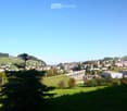 A panoramic view of a town with a mix of residential and commercial buildings, surrounded by green hills and a clear blue sky. A prominent aqueduct structure is visible in the distance.