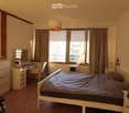 Cozy bedroom with a double bed, wooden floor, large window with curtains, desk with chair, mirror, and nightstand.