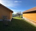 A grassy yard between two yellow buildings with tiled roofs, surrounded by a rural landscape with hills and a clear blue sky.