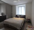A modern bedroom with a large bed, nightstand, and a small table with a book on it. The room features wooden flooring, large windows with blinds, and a dark-colored wardrobe.