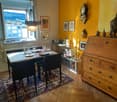 A cozy dining area with a table set for four, surrounded by chairs, a wooden cabinet, and decorative items including a clock and framed pictures. The room features warm yellow walls, wooden flooring, and a large window offering a scenic view.