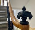 Close-up of a wooden handrail with ornate black metal balusters and a decorative finial, part of a staircase in a well-lit interior space.