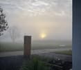 Garden area with gravel, small plants, wooden post, and a view of a misty field with trees and a setting sun.