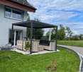 Outdoor patio area with a black metal pergola, wooden furniture including a sofa, table, and chairs, and potted plants. The area is surrounded by a well-maintained lawn and a modern house with large windows and a solar panel on the roof.