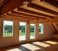 Bright, empty attic space with wooden beams and exposed ceiling, large windows with views of greenery, and wooden flooring.