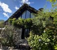 A rustic stone house with a unique roof and a small entrance, surrounded by lush greenery and flowers.
