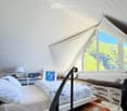 Bright bedroom with a large bed, nightstand, lamp, books, and a painting, featuring large windows with a scenic view.