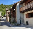 A row of traditional wooden and stone houses in a narrow village street, surrounded by greenery and mountains.