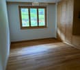 Empty room with wooden flooring, light-colored walls, and a large window with wooden frame and blinds. A built-in wooden wardrobe is visible on the right side.