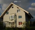 Two-story house with light gray exterior, large windows, and a modern design. Features include a prominent entrance with glass doors, a small balcony, and decorative sunflowers in the front yard.