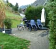 Outdoor patio with a table, chairs, umbrella, grill, and potted plants, surrounded by greenery and a wooden fence.
