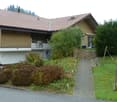 Two-story house with a tiled roof, wooden siding, and a balcony. The house is surrounded by greenery, including bushes, trees, and a small garden area with a wooden fence and a pathway leading to the entrance.