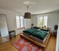 Modern bedroom with wooden floor, large bed with green duvet, colorful rug, mirror, small stool, table lamp, potted plants, and books on nightstand.