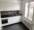Modern kitchen with white cabinets, black countertops, stainless steel sink, and built-in oven. Dark tiled floor and radiator visible.
