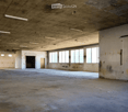 Empty industrial-style room with concrete floors and walls, large windows, and exposed ceiling with fluorescent lighting.