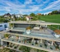 Modern two-story house with a large terrace and glass railings, surrounded by greenery and potted plants. The house features a flat roof and contemporary design.