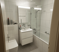 Modern bathroom with white tiles, a toilet, a sink with a mirror, a bathtub with a glass shower stall, and a light fixture above the mirror.