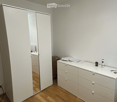A furnished room with a large white wardrobe, a white drawer unit, and a mirror. The room features wooden flooring and white walls.