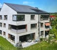 Modern two-story house with grey concrete facade, multiple windows, balconies, and a well-maintained garden area with outdoor furniture.