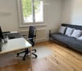 A modern, multi-functional room with a grey sofa, a black office chair, a white desk, and a radiator. The room features large windows with a view of greenery outside.